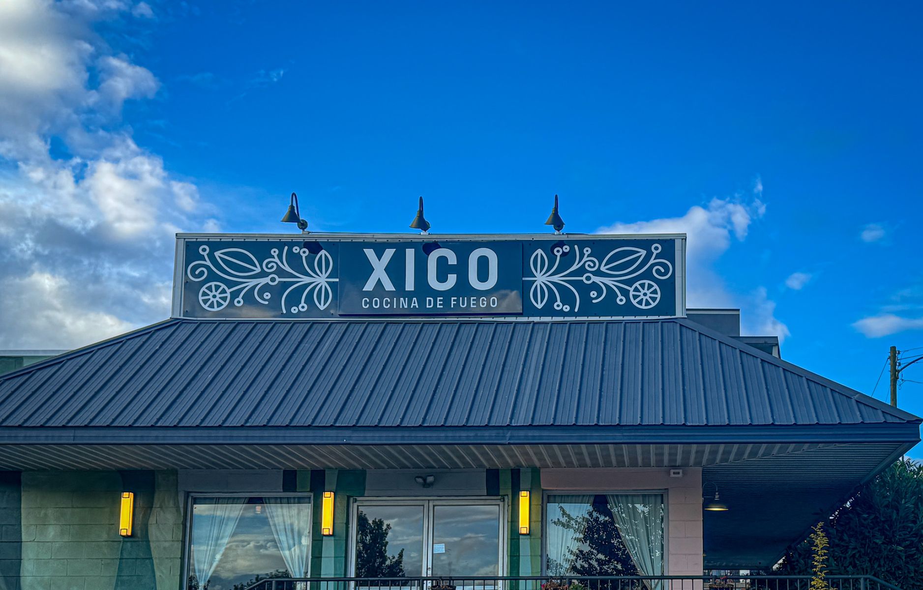 Restaurant sign reading 'XICO' with decorative white flourishes on blue, above the entrance of a building against a clear blue sky.