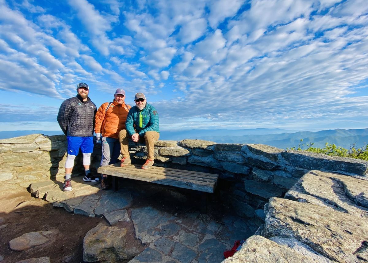 Three hikers at the summit of Craggy Pinnacle with sweeping views of the Blue Ridge Mountains under a brilliant blue, cloud-filled sky.