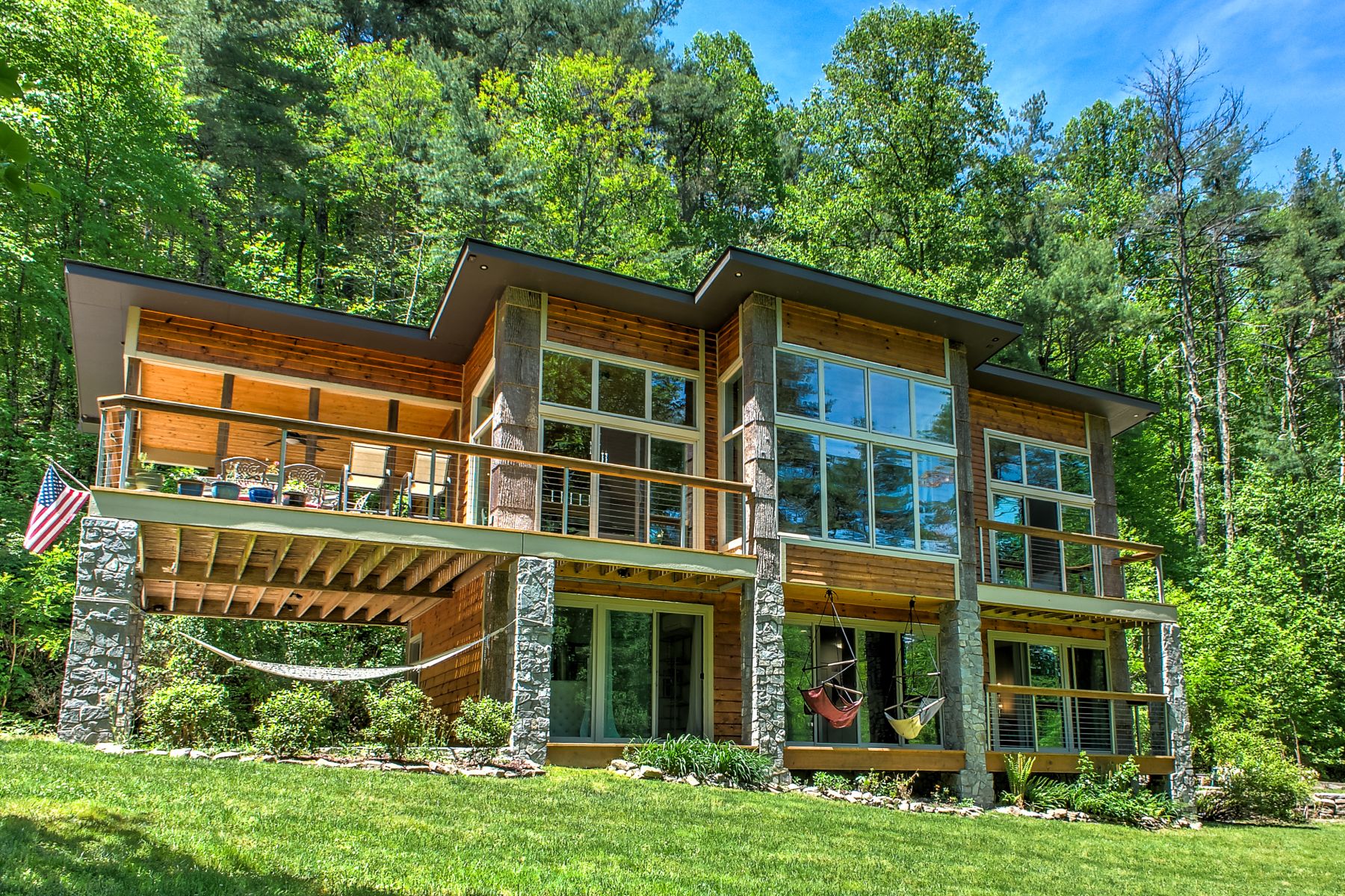 Exterior of Vanaprastha, a modern mountain home featuring natural wood siding, stone columns, expansive windows, and wraparound decks surrounded by lush forest.
