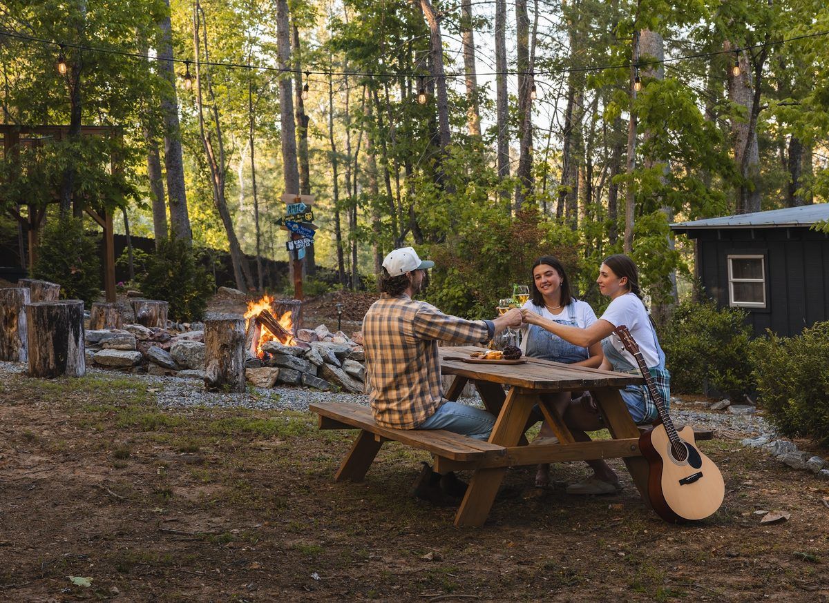 Three guests sharing a toast at an outdoor picnic table at The A-Frame Nest, surrounded by trees, string lights, a glowing fire pit, and an acoustic guitar.