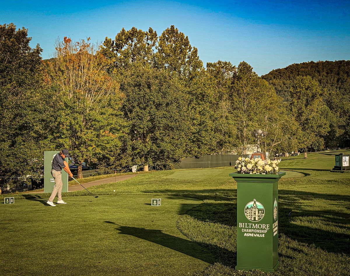 Professional golfer tees off at the 2026 Biltmore Championship Asheville at The Cliffs at Walnut Cove, with the championship trophy and Blue Ridge Mountains in the background.