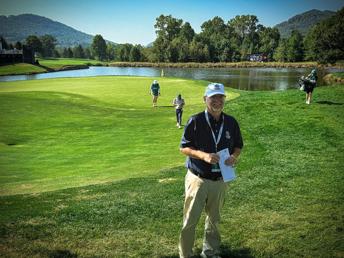 Biltmore Championship Asheville volunteer stands beside a putting green overlooking a scenic pond and the Blue Ridge Mountains at The Cliffs at Walnut Cove during the 2026 PGA TOUR event.