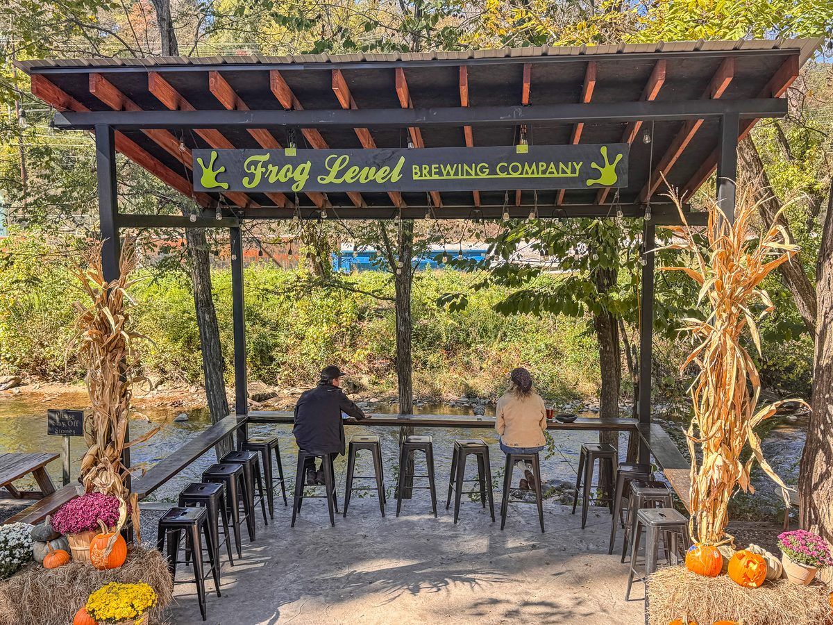 Outdoor creekside seating at Frog Level Brewing Company in Waynesville, North Carolina, featuring a covered pavilion, bar stools, guests relaxing by the water, and festive autumn decorations.